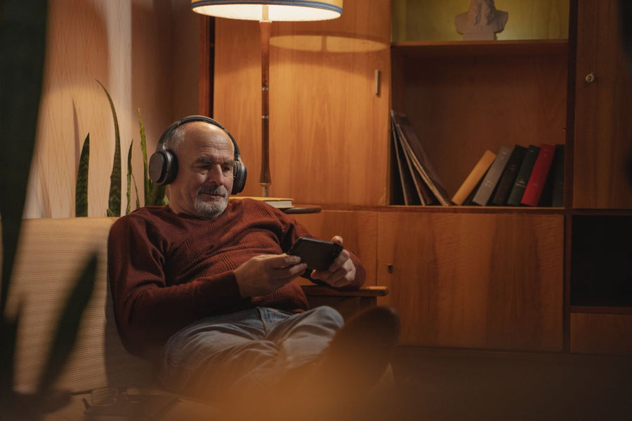 Older man listening through headphones while holding a phone in a quiet room.